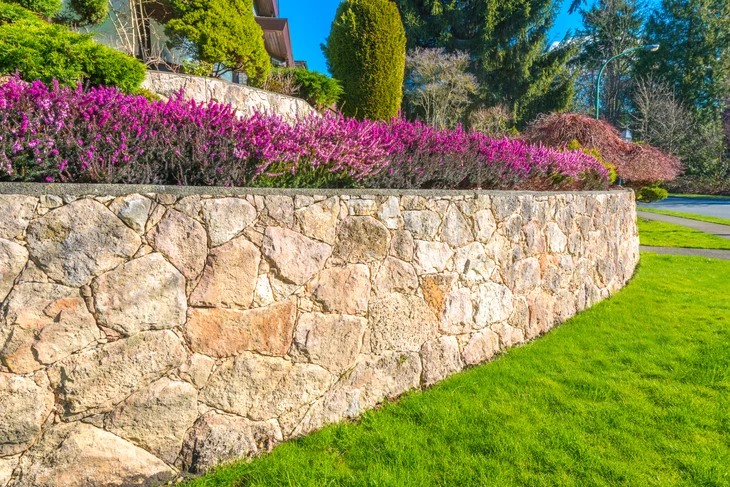 Aménagement extérieur en terrasses à niveaux multiples sur terrain en pente. Au premier plan, escalier droit en granit gris flammé avec environ 8-10 marches bien définies, bordures latérales en pierre naturelle. Chaque niveau de terrasse accueille une plage plane en dallage de granit ou pierre naturelle, délimitée par des murets bas en maçonnerie paysagère (pierre ou béton enduit). Végétation arbustive et massifs paysagers intégrés entre les niveaux, créant une composition harmonieuse. Pelouse ou revêtement de sol en gravillon entre certaines terrasses. Maison d'habitation visible en arrière-plan, ciel dégagé. Photo professionnelle en plein jour, mettant en valeur la qualité des matériaux et la structure en niveaux du terrain.