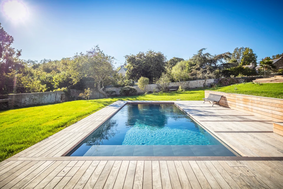 Piscine rectangulaire extérieure avec liner bleu clair, entourée d'une terrasse harmonieuse en dalles de granit gris flammé. Margelles en pierre naturelle beige, pool-house moderne en arrière-plan, jardin paysager avec arbustes et topiaires. Enrouleur de bâche solaire visible. Environnement soigné et cohérent, mettant en évidence l'harmonie entre la piscine et son aménagement extérieur.